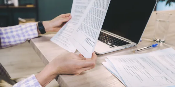 Mãos entregando um documento impresso sobre uma mesa com notebook, papéis e caneta.