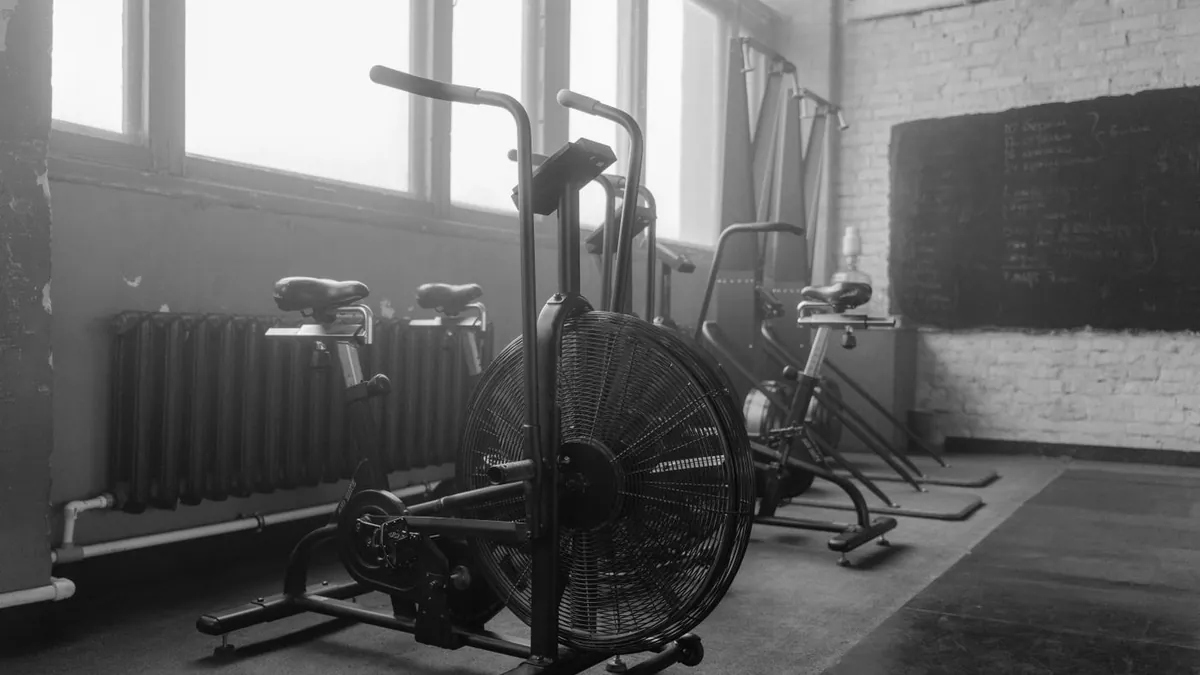 Bicicletas de treino e equipamentos alinhados no salão da academia, em preto e branco