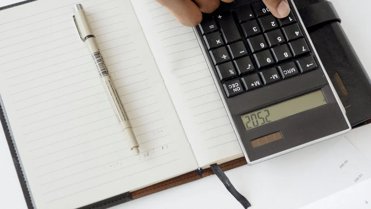 Mão usando uma calculadora ao lado de um caderno aberto e de um relatório