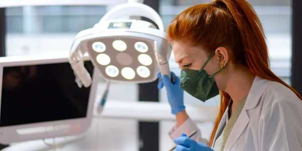 Dentista de jaleco e máscara atendendo uma paciente na cadeira, sob o foco do refletor