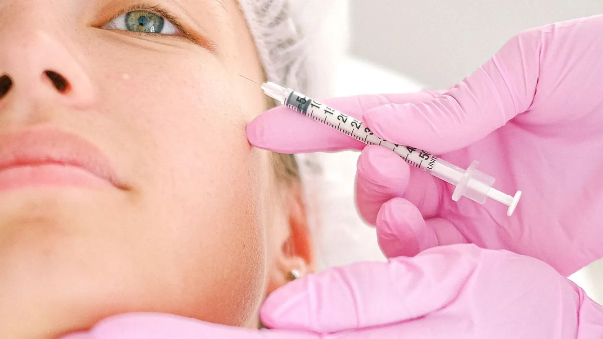 Cliente de touca deitada recebendo aplicação facial de uma profissional de luvas rosa
