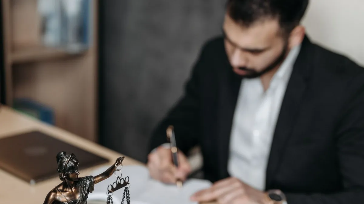 Homem de terno assinando um documento na mesa, com a estátua da Justiça em primeiro plano