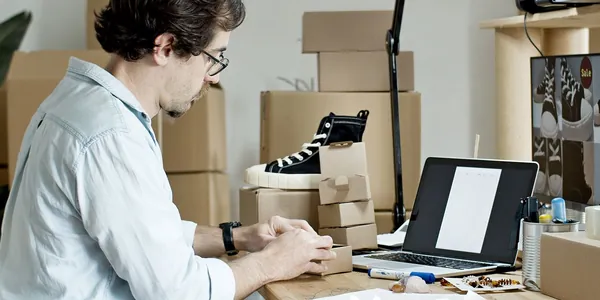 Homem embalando pedidos em caixas de papelão com o notebook aberto na bancada
