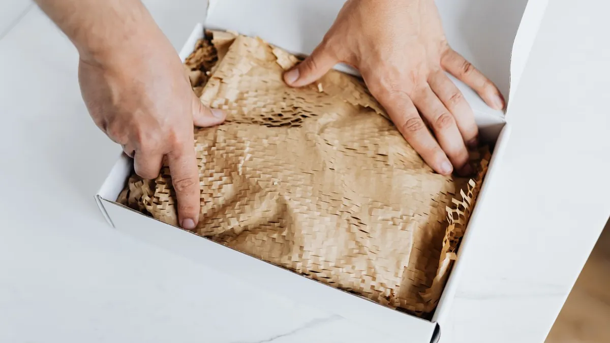 Mãos acomodando papel kraft dentro de uma caixa branca de envio