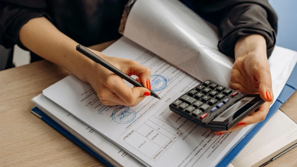 Mão escrevendo num caderno ao lado da calculadora e de uma pilha de documentos