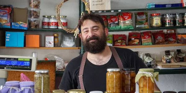 Lojista sorrindo atrás do balcão de uma loja de produtos naturais, com prateleiras cheias ao fundo.