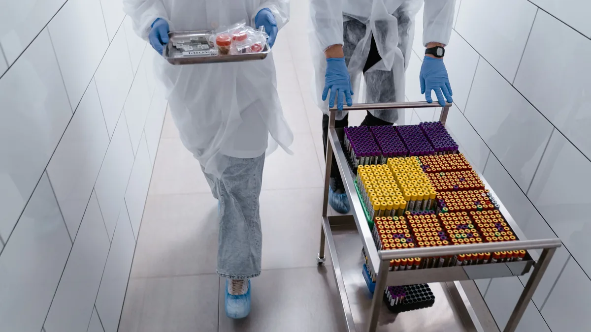Lab workers in protective gear moving sample trays in a sterile laboratory hallway.