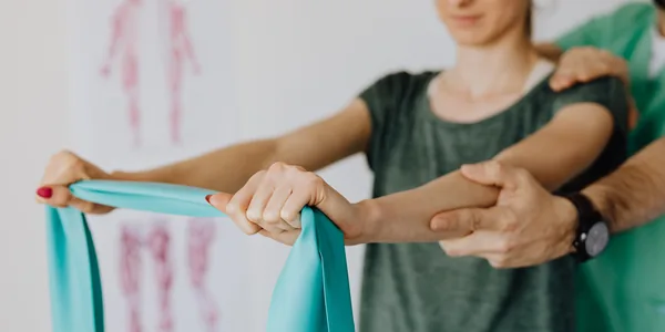 Crop anonymous orthopedist in wristwatch and uniform helping young woman in casual wear reaching arms with elastic tape in doctor office