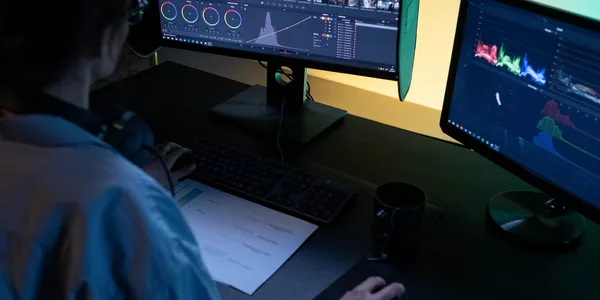 A video editor using dual monitors for editing footage in a dimly lit studio.