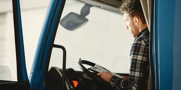 Motorista dentro da cabine de um caminh&atilde;o azul conferindo um documento apoiado no volante