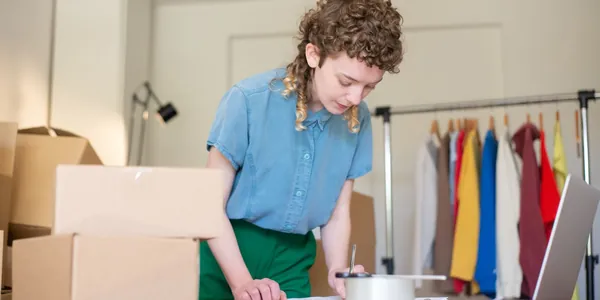 Empreendedora anota pedidos sobre a mesa da própria loja, entre caixas de papelão, notebook e arara de roupas coloridas, situação comum de quem descobre que o MEI estourou o limite de faturamento