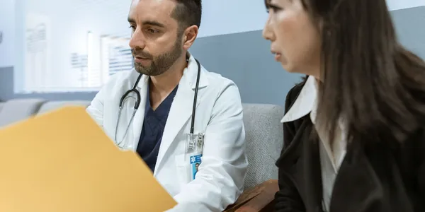 M&eacute;dica de jaleco branco sentada &agrave; mesa do consult&oacute;rio conferindo relat&oacute;rios em um notebook