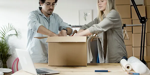 Duas pessoas embalando pedidos de loja virtual em uma caixa, com notebook aberto na mesa