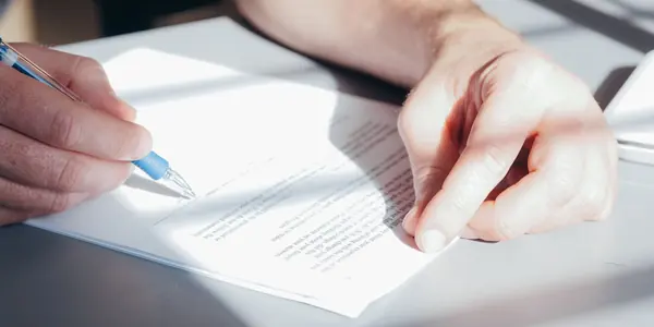 Mão escrevendo em um documento sobre a mesa, representando a redação do objeto social no contrato da empresa