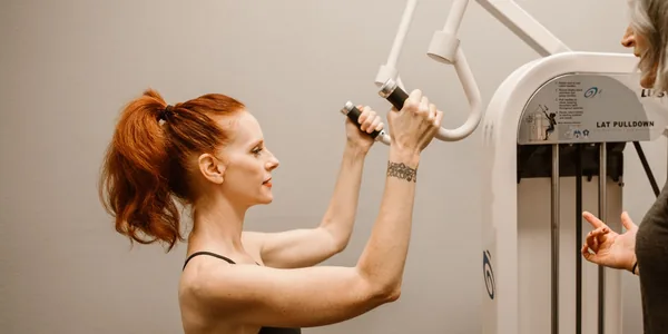 Aluna treinando em um aparelho da academia com orientação do instrutor