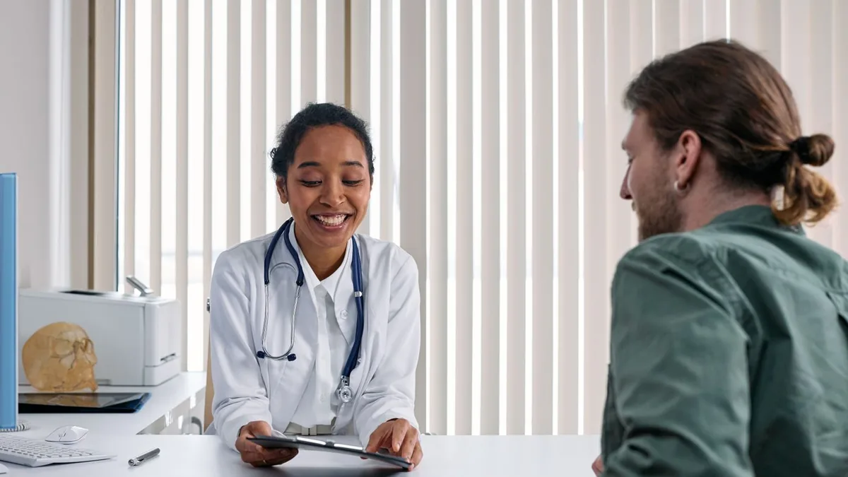 Médica de jaleco branco conversando e sorrindo com um paciente no consultório