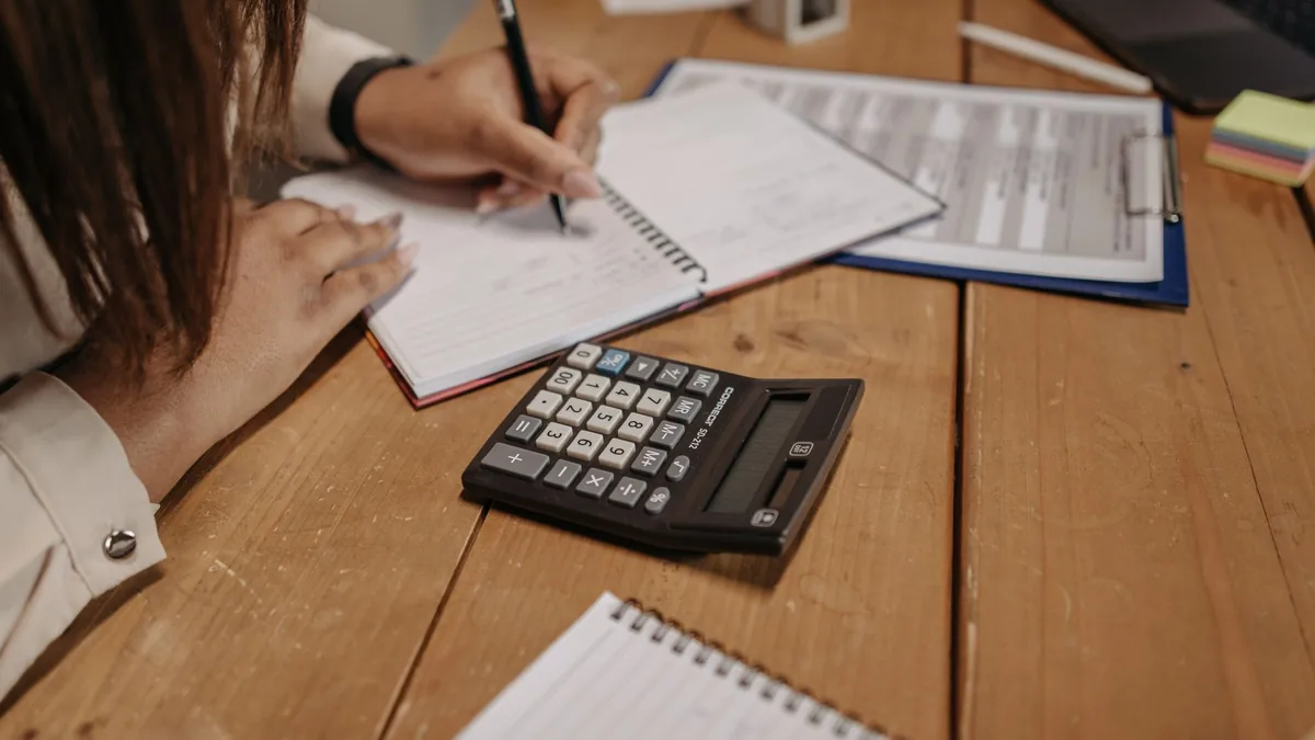 Mulher anotando números ao lado de uma calculadora sobre a mesa de madeira