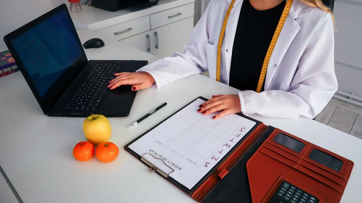 Nutricionista com notebook, prancheta e frutas sobre a mesa do consult&oacute;rio