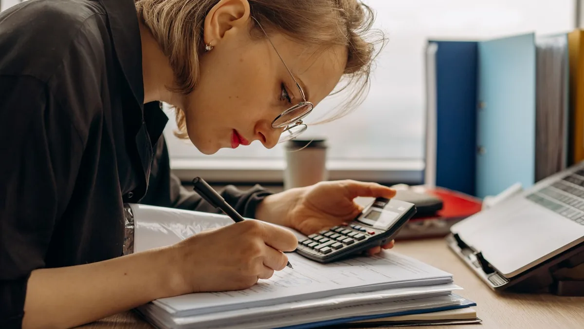 Mulher fazendo contas com calculadora e documentos sobre a mesa
