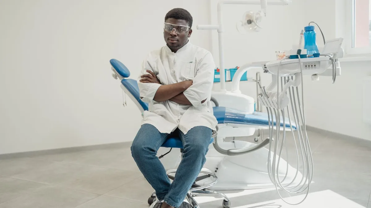 Dentista de jaleco sentado ao lado da cadeira do consultório