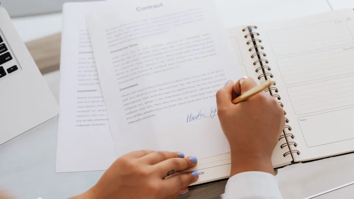 Mão assinando um contrato ao lado de um caderno e um notebook