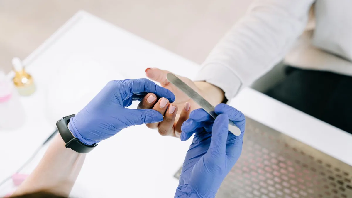 Profissional de luvas azuis lixando a unha de uma cliente na mesa de manicure