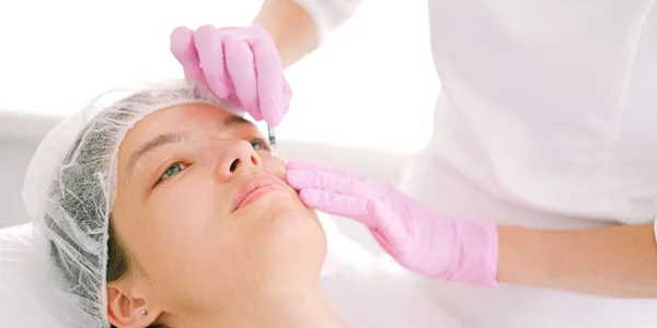Profissional de luvas cor-de-rosa aplicando um procedimento estético no rosto de uma cliente deitada.