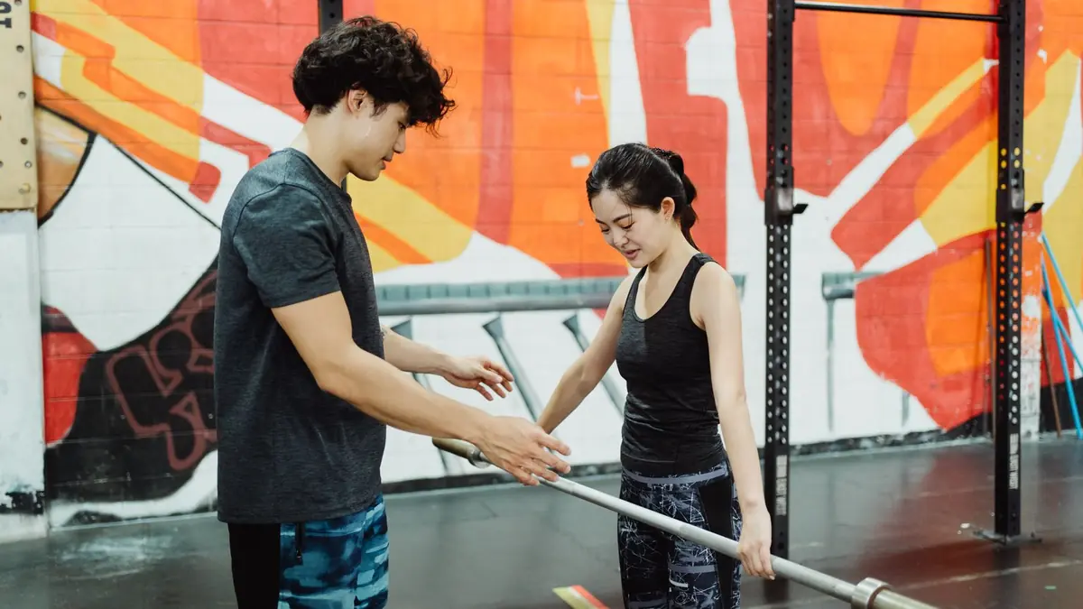 Instrutor orientando uma aluna em exercício com barra dentro da academia.