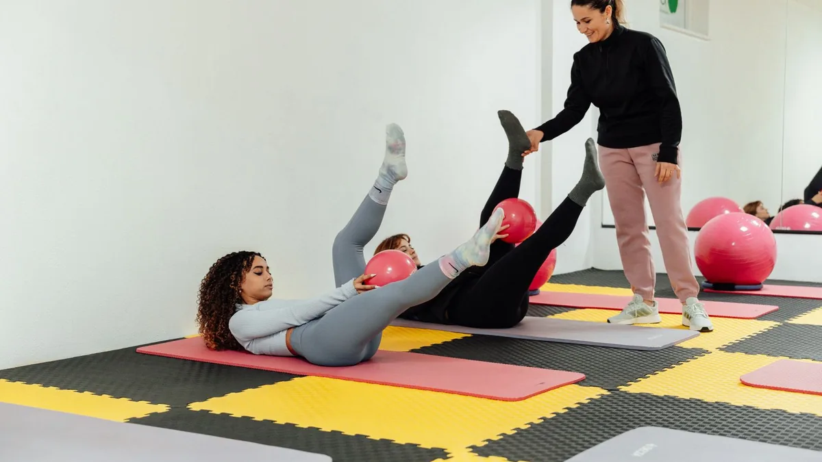 Professora auxiliando uma aluna em exercício de solo sobre o tatame.