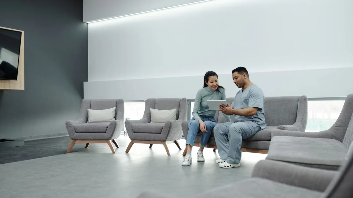 Sala de espera clara de uma clínica, com poltronas e duas pessoas sentadas ao fundo.
