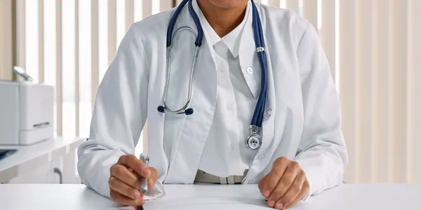 Médica de jaleco branco com estetoscópio sentada à mesa do consultório.