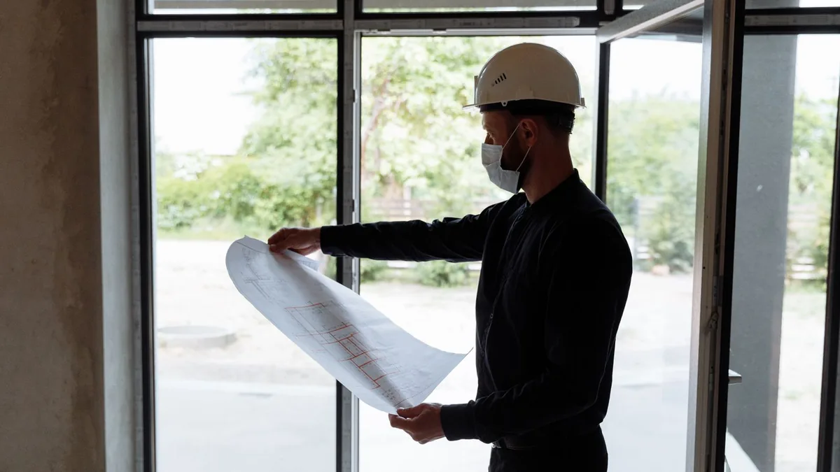 Profissional com capacete segurando uma planta impressa junto à janela da obra.