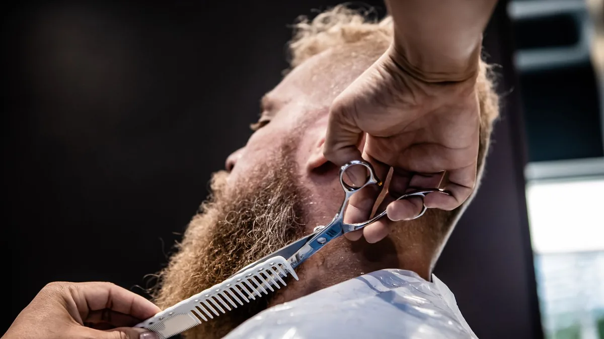 Barbeiro aparando a barba de um cliente com pente e tesoura.