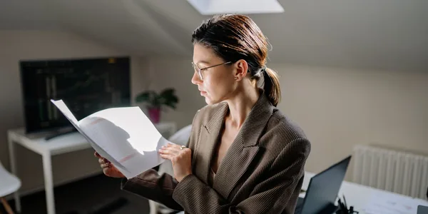 Mulher de blazer xadrez marrom lendo um relat&oacute;rio impresso em p&eacute; no escrit&oacute;rio, com um monitor de gr&aacute;ficos ao fundo