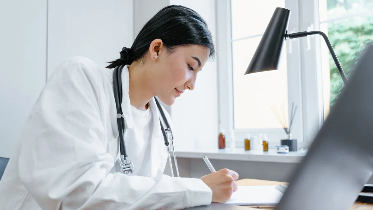 Médica escrevendo anotações na mesa do consultório, ao lado do computador