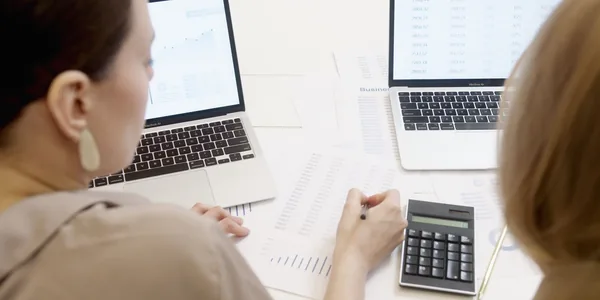 Duas pessoas analisando gráficos e dados financeiros em notebooks, representando simulações da Reforma Tributária