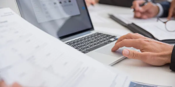 Mãos sobre o teclado de um notebook ao lado de documentos impressos em uma mesa de reunião