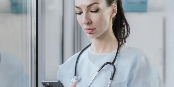 Profissional de sa&uacute;de de jaleco e estetosc&oacute;pio usando o celular no consult&oacute;rio