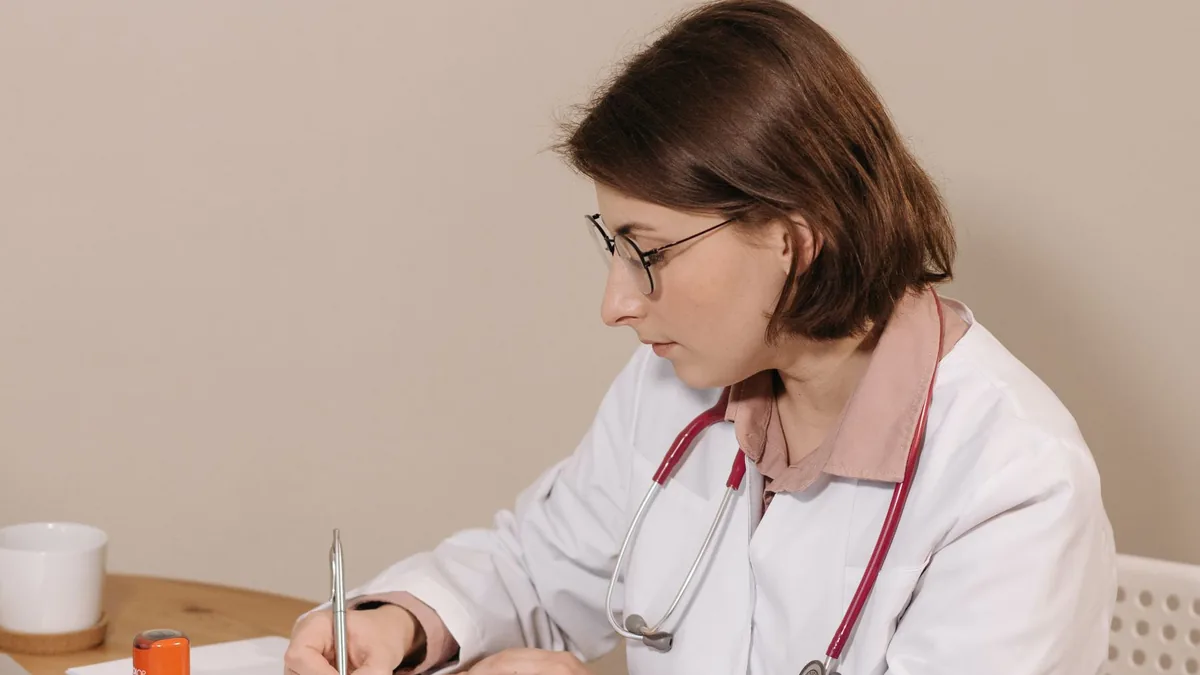 Médica com estetoscópio anotando em um caderno na mesa do consultório