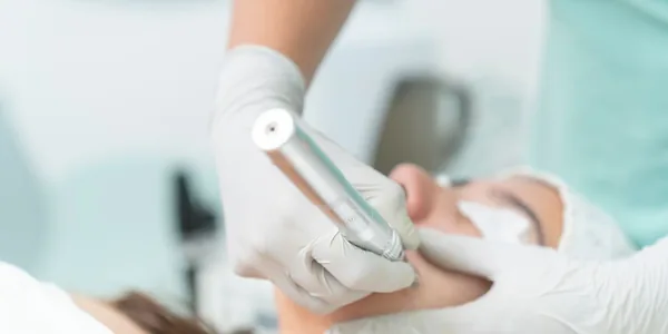 Profissional de luvas aplicando um aparelho de tratamento no rosto de uma cliente deitada na maca da cl&iacute;nica de est&eacute;tica