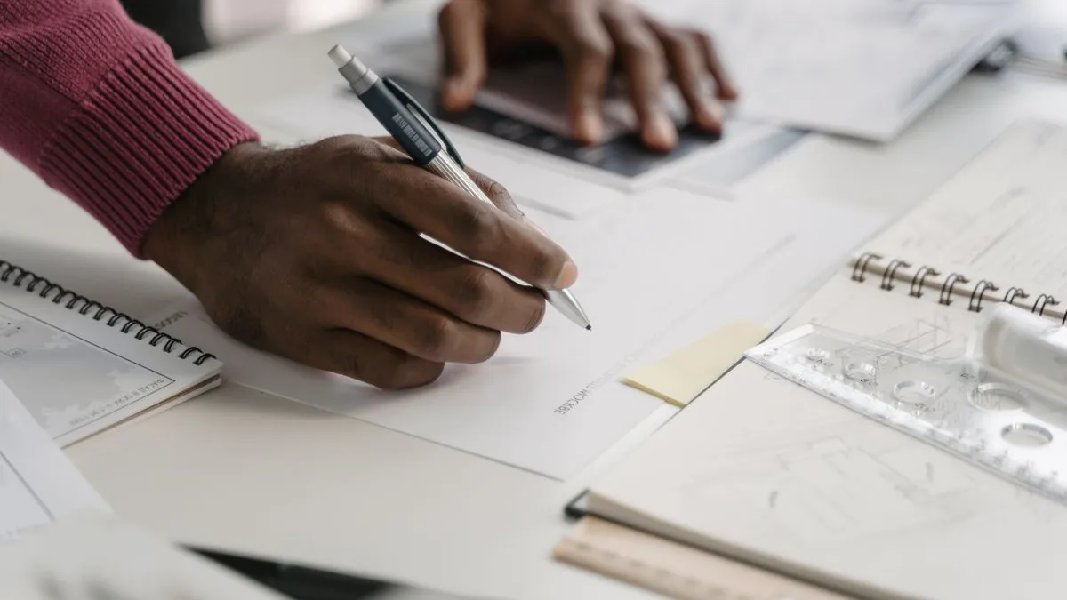 M&atilde;os de um profissional desenhando sobre a planta do projeto com l&aacute;pis e r&eacute;gua, ao lado do notebook e de cadernos t&eacute;cnicos