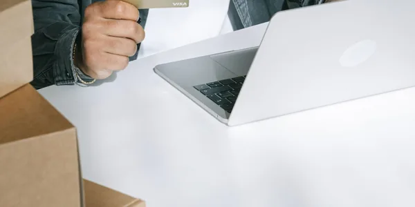 Mão fechando uma caixa de papelão ao lado de um notebook aberto sobre a mesa.