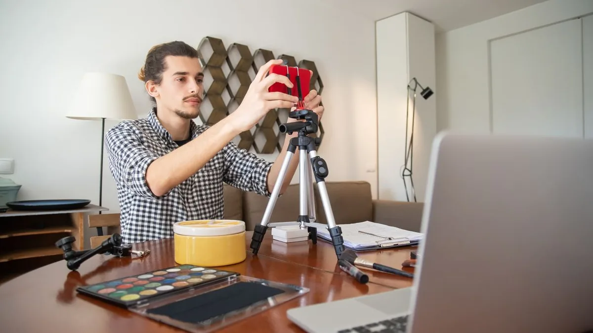 Criador de conte&uacute;do ajustando o celular no trip&eacute; antes de gravar, com notebook e materiais espalhados na mesa
