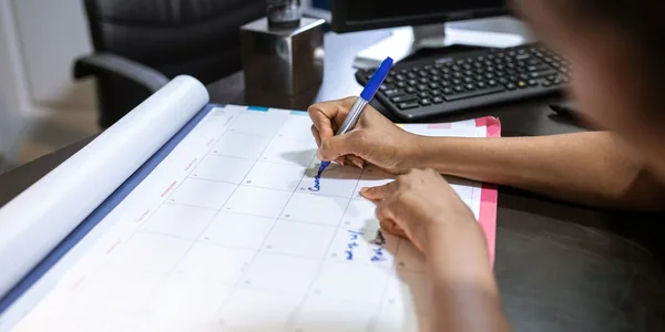 M&atilde;os de uma mulher marcando uma data com caneta azul num calend&aacute;rio de mesa, ao lado do teclado do computador
