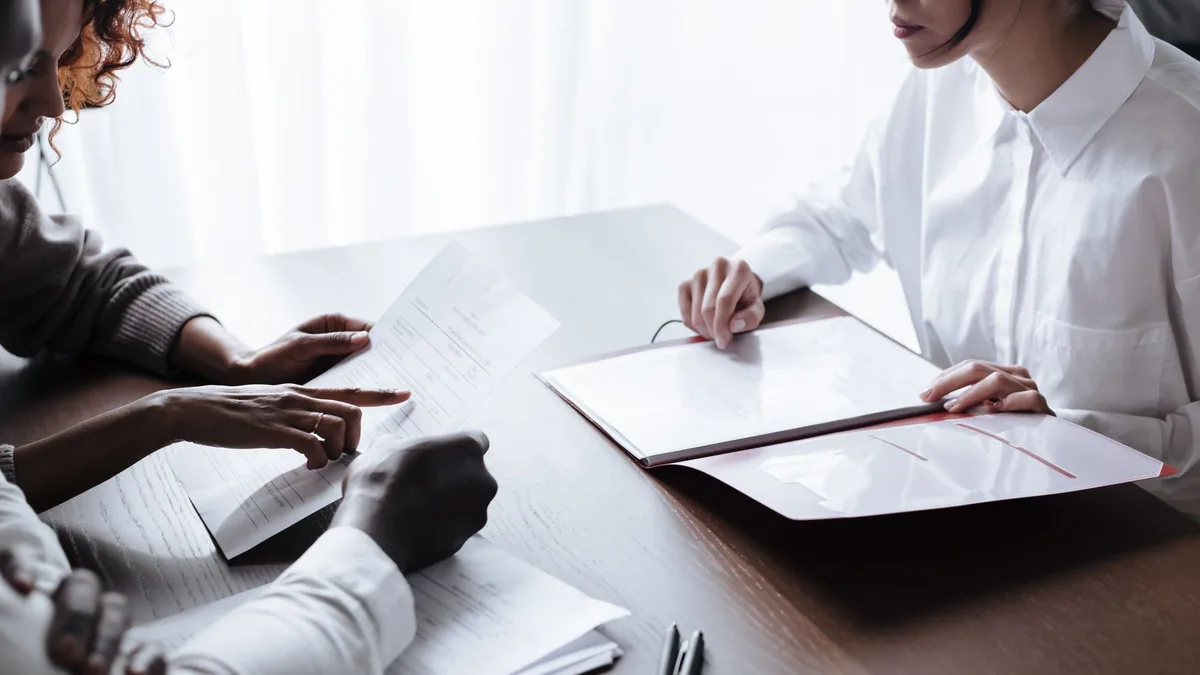 Tr&ecirc;s pessoas sentadas &agrave; mesa conferindo papeis e uma pasta aberta durante uma reuni&atilde;o de escrit&oacute;rio