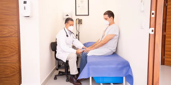 Médico de jaleco branco e estetoscópio aferindo a pressão de um paciente sentado na maca de um consultório.