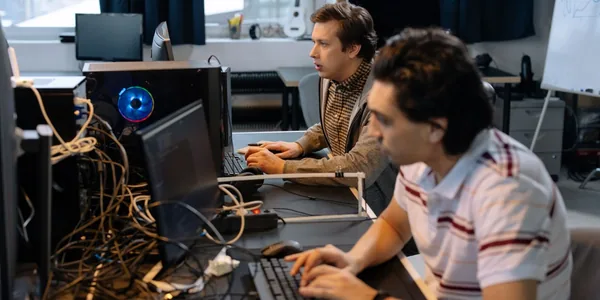Dois desenvolvedores trabalhando lado a lado em monitores de uma bancada de escrit&oacute;rio, com cabos e computadores ao redor
