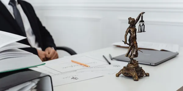 Mesa de escritório de advocacia com estatueta da deusa da justiça, documentos, caneta e notebook, e um profissional de terno ao fundo.