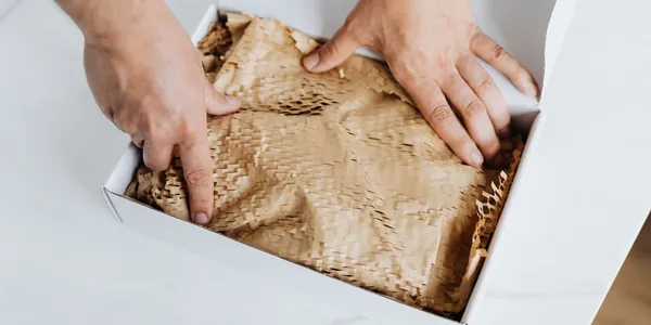 Mãos ajeitando papel kraft de proteção dentro de uma caixa branca de envio, sobre uma mesa clara.