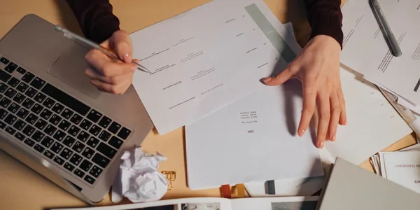 Pessoa fazendo anotações no notebook cercada de documentos sobre a mesa